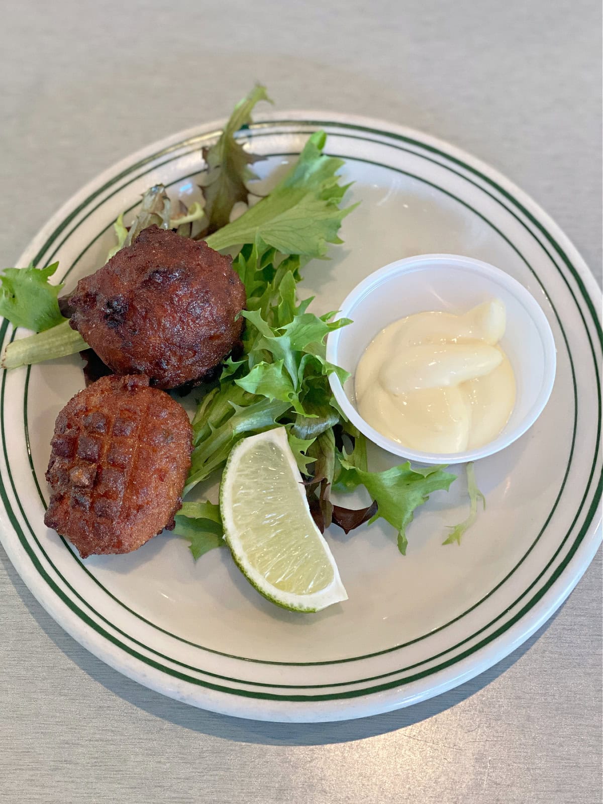 Two conch fritters on a plate with lettuce, lime, and sauce.