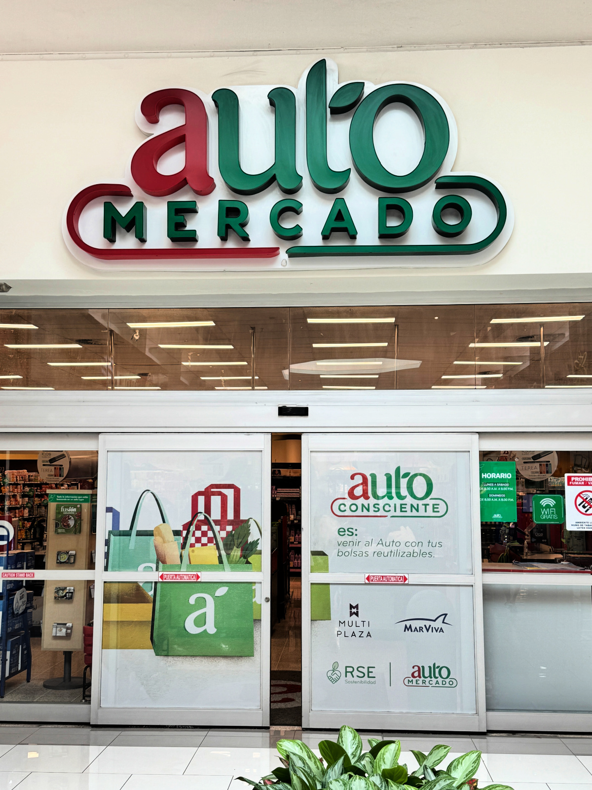 Entrance of an Auto Mercado grocery store in Costa Rica, featuring the red and green Auto Mercado logo above glass sliding doors with reusable bag signage.