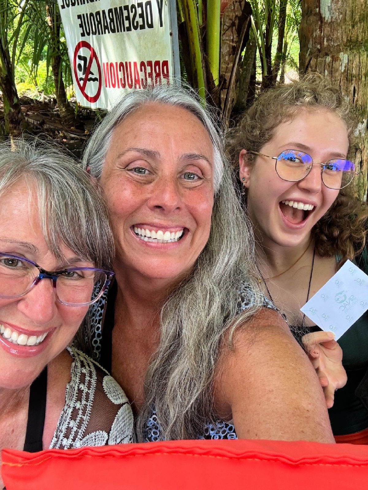 Three smiling women taking a cheerful selfie in a tropical Costa Rican setting with palm trees and warning signs in the background.