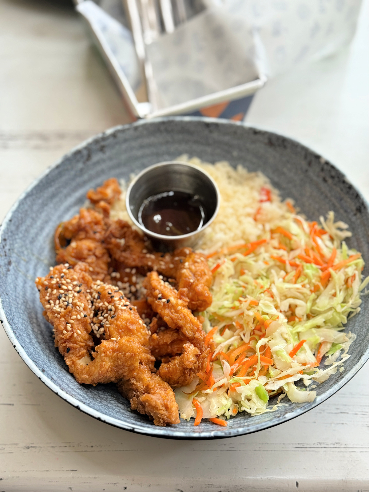 A gray ceramic bowl filled with crispy sesame-topped fried chicken strips, shredded cabbage slaw, and a serving of rice, with a small metal cup of dark dipping sauce in the center.