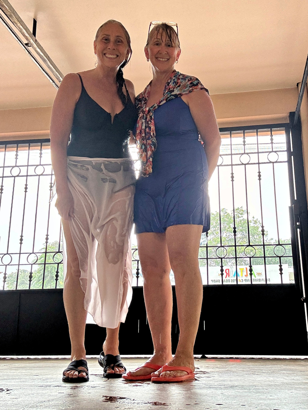 Two smiling women standing indoors, soaked from the rain, wearing swimsuits and flip-flops with a wet patio floor beneath them and an iron gate in the background.