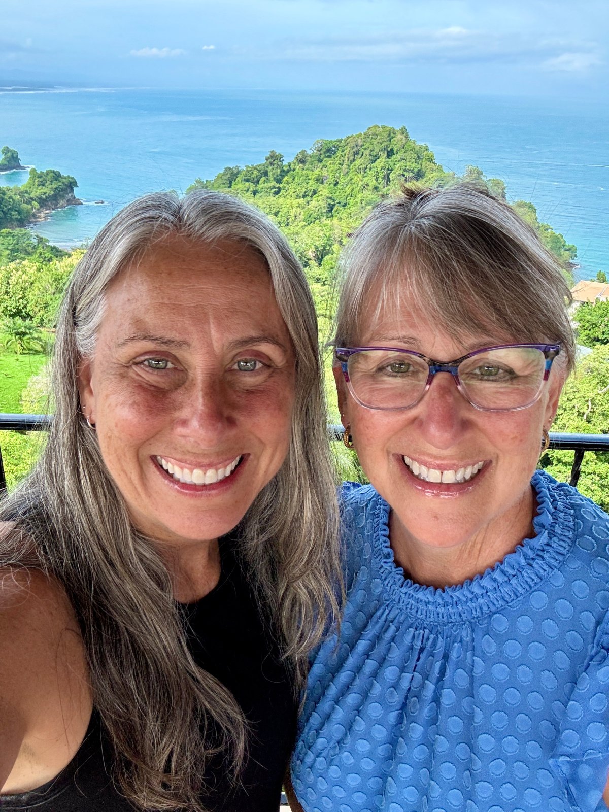 Two smiling women posing in front of a scenic overlook with lush green cliffs and the Pacific Ocean in the background in Costa Rica.