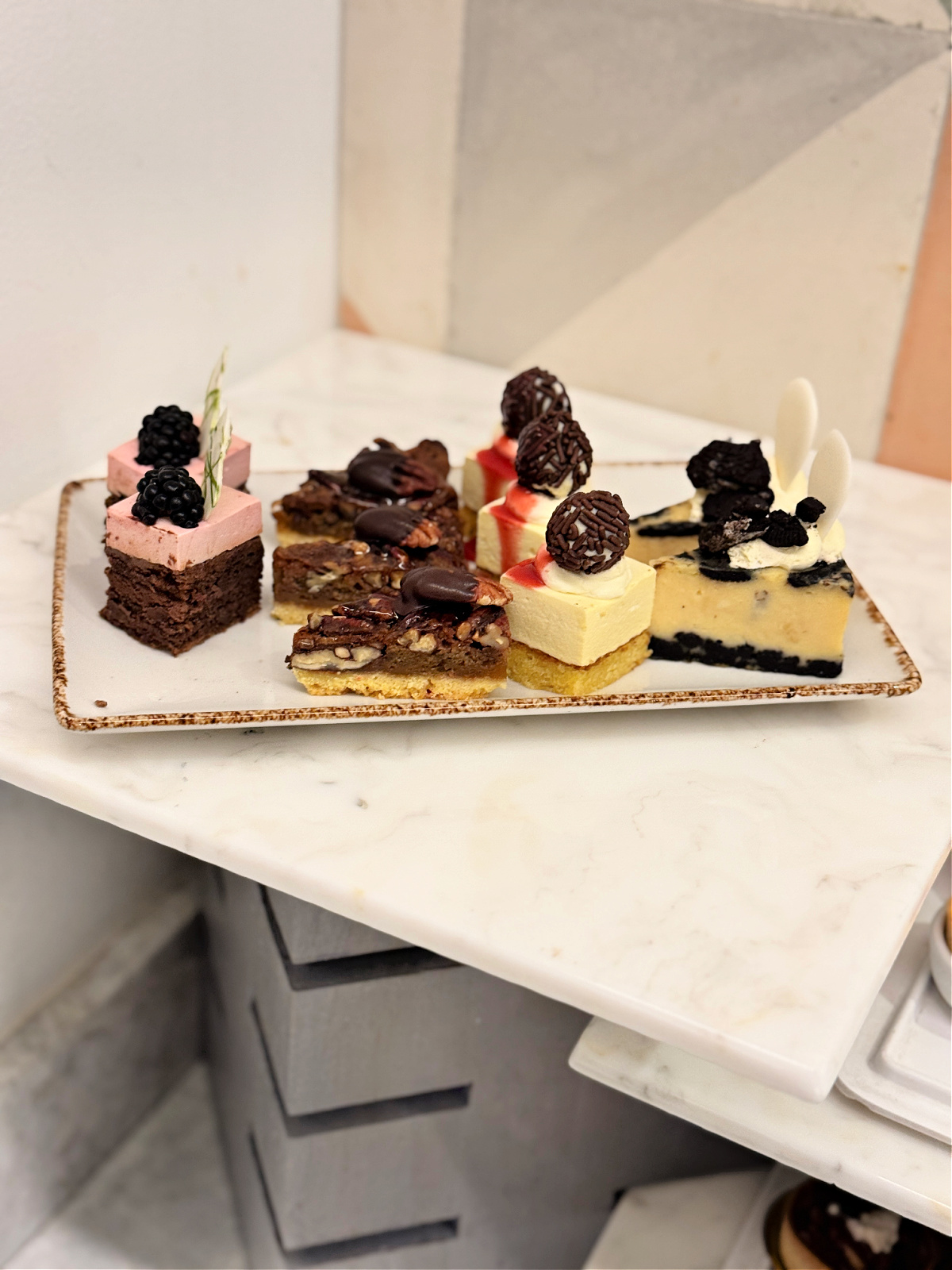A rectangular platter of assorted mini desserts, including berry mousse bites, pecan chocolate bars, chocolate truffle-topped cakes, and cookies-and-cream cheesecake squares, displayed on a marble countertop.