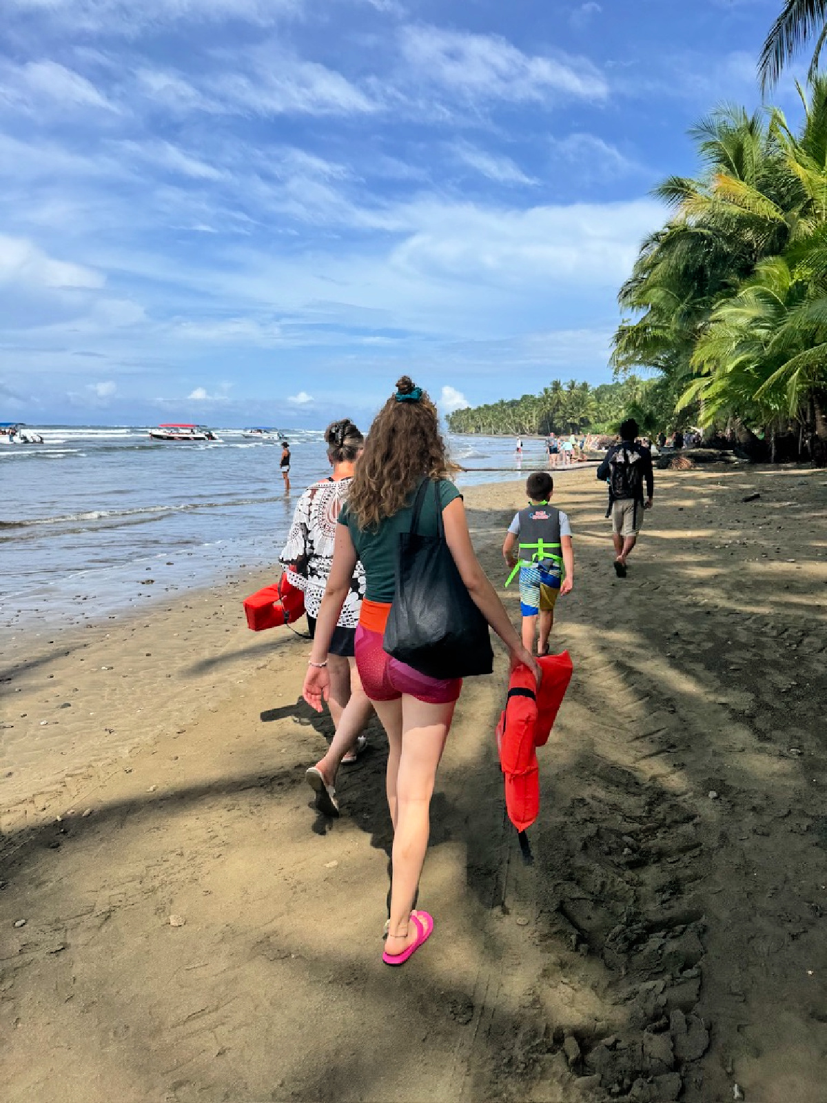 A small group walking along a tropical beach toward the water, carrying bright red flotation devices, with palm trees and boats lining the shoreline.