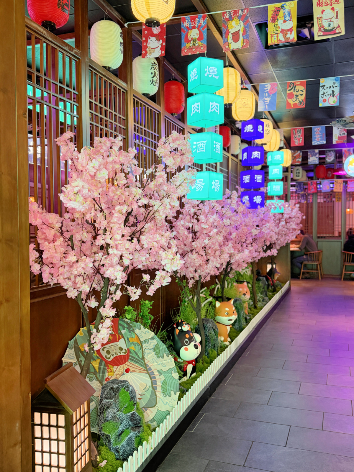 Colorful Japanese restaurant interior with cherry blossom decor, hanging lanterns, illuminated signs, and decorative figurines lining a dining corridor.