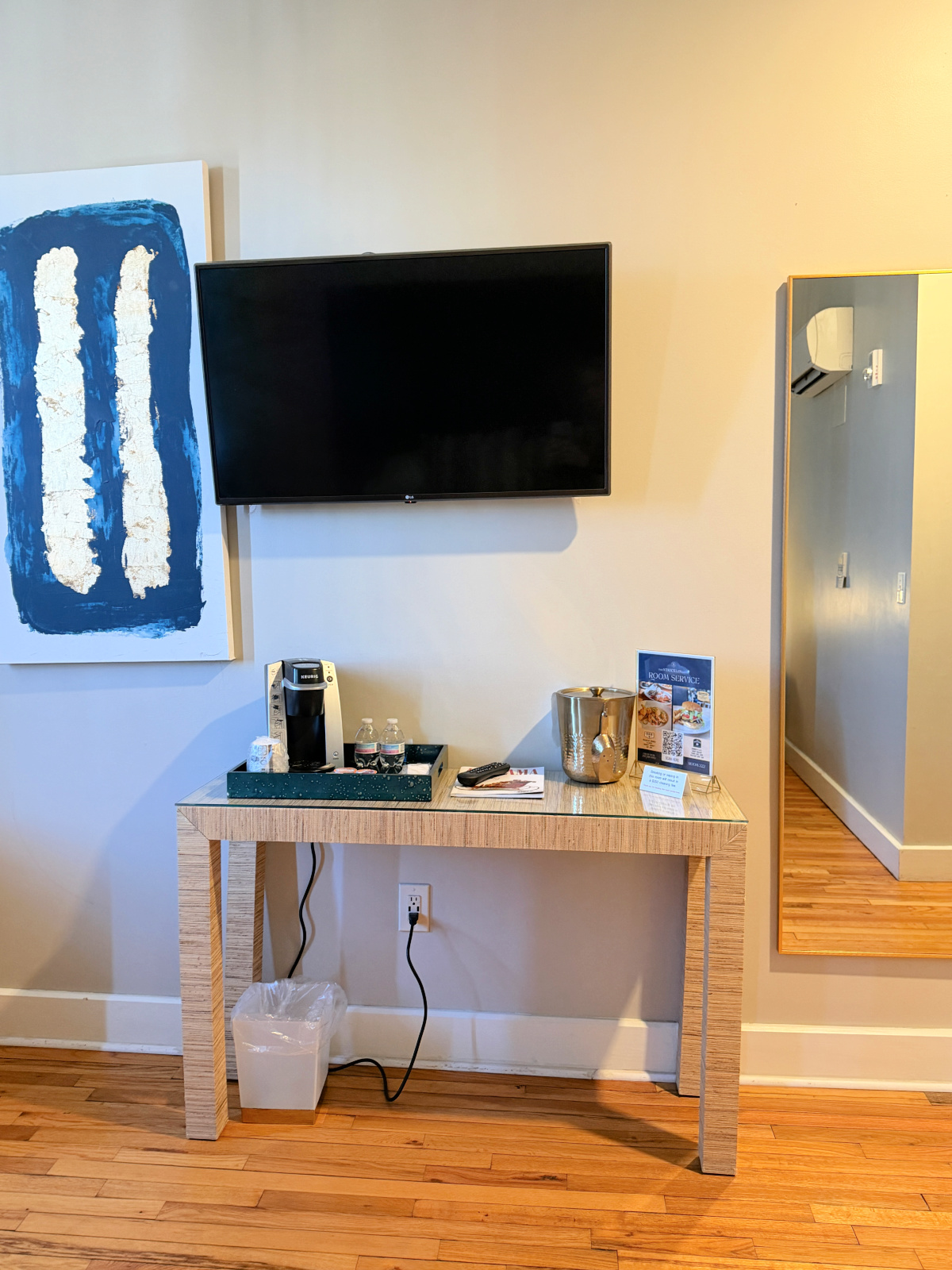 In-room coffee station and wall-mounted TV in a guest room at The Stricklin Hotel in Florence, Alabama, with Keurig coffee maker, bottled water, and modern artwork.
