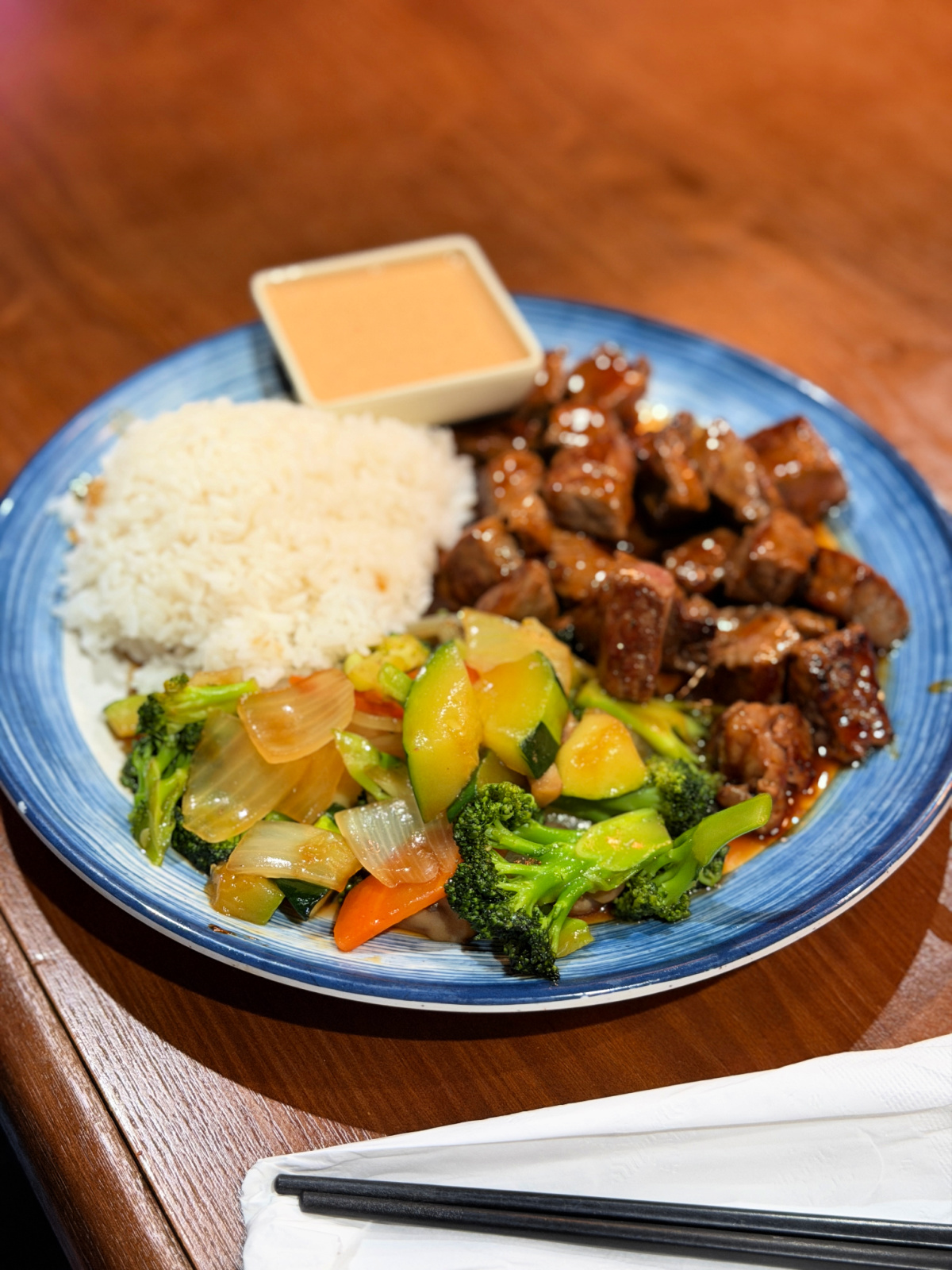 Teriyaki steak bites served with white rice, stir-fried vegetables, and dipping sauce on a blue plate at a Japanese restaurant.