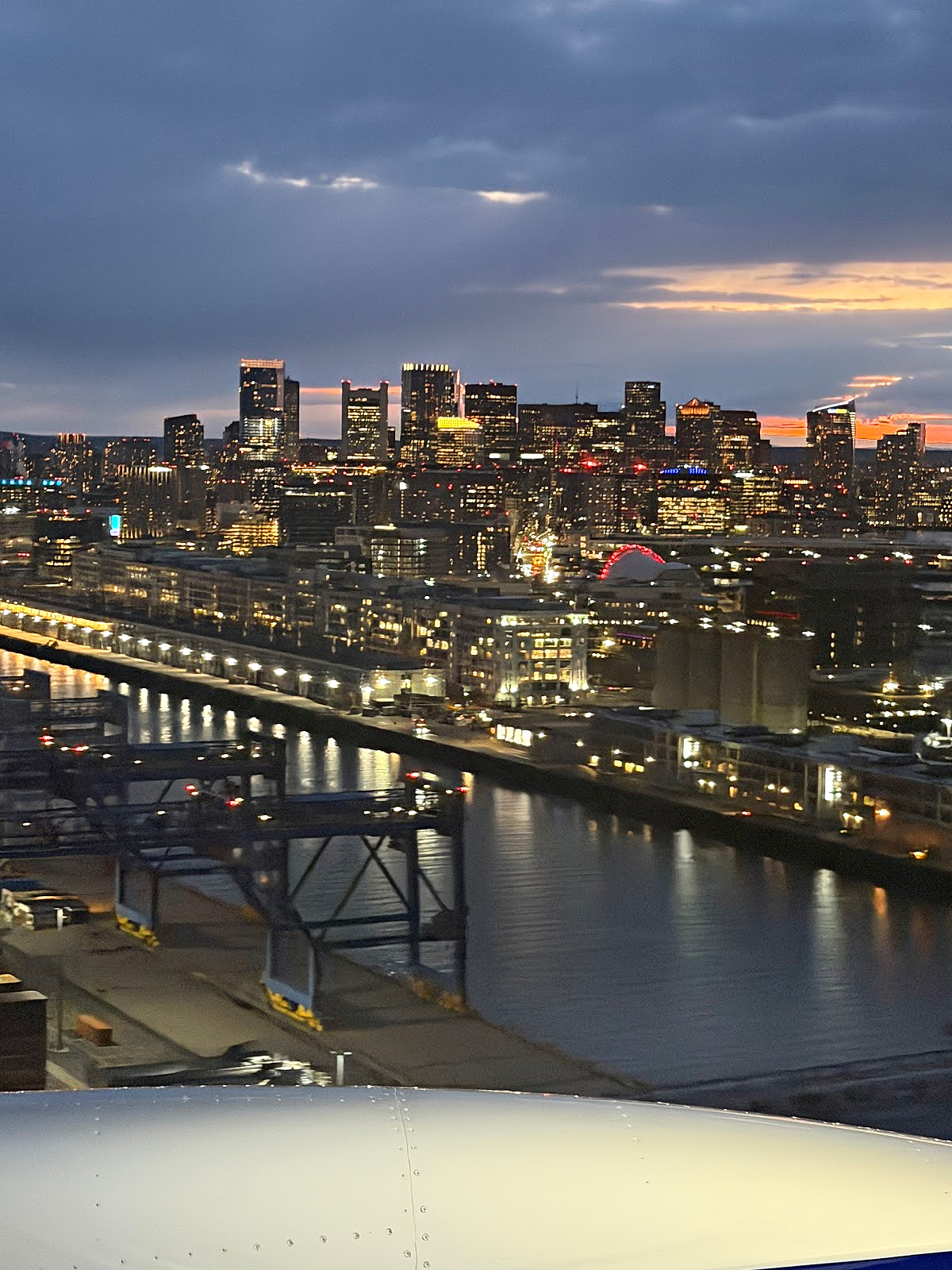 Boston skyline glowing at sunset as seen from a JetBlue airplane window during departure, with city lights reflecting on the water below.