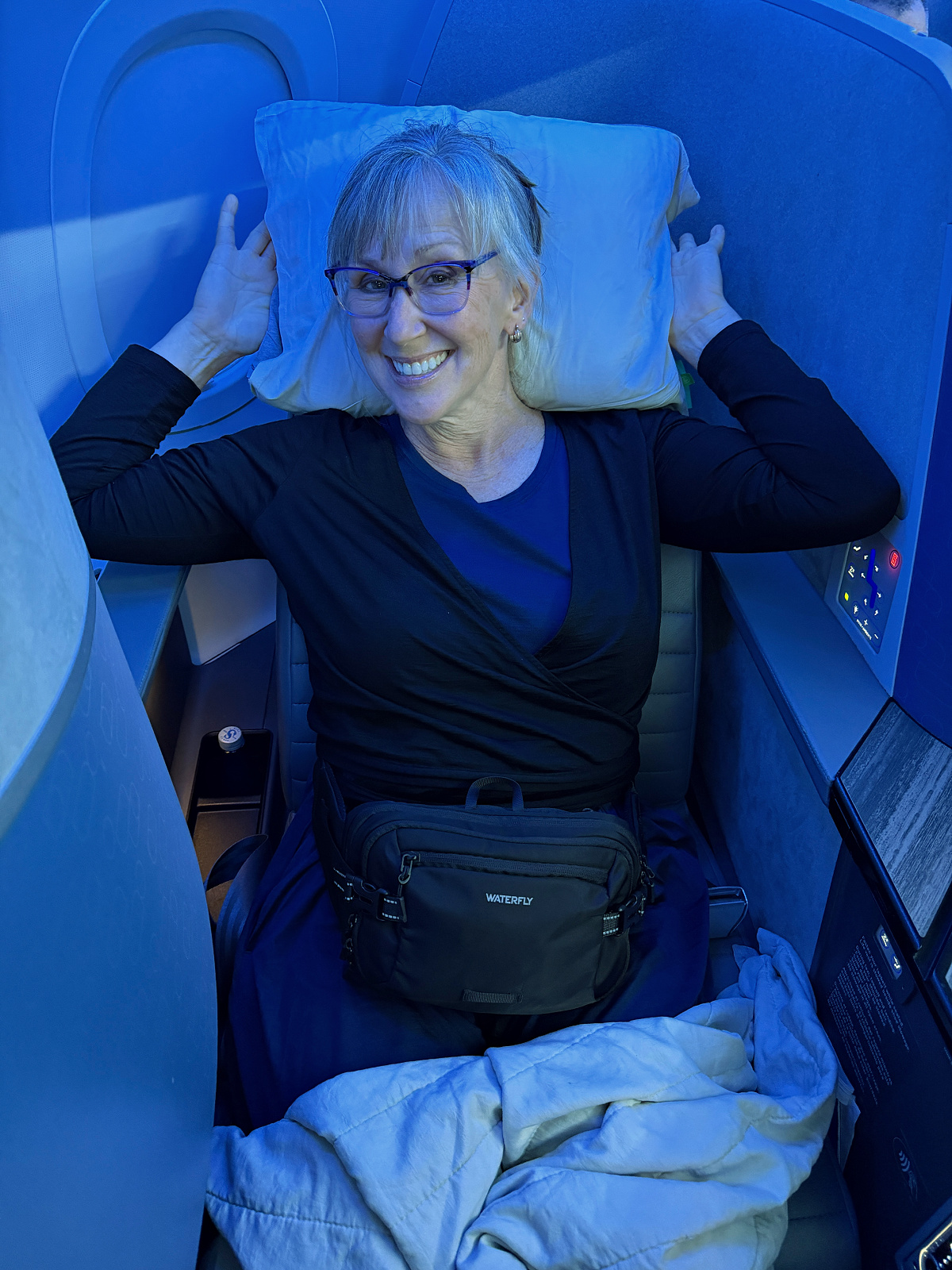 Passenger smiling while relaxing in a JetBlue Mint lie-flat seat with pillow and blanket in a private Mint suite.