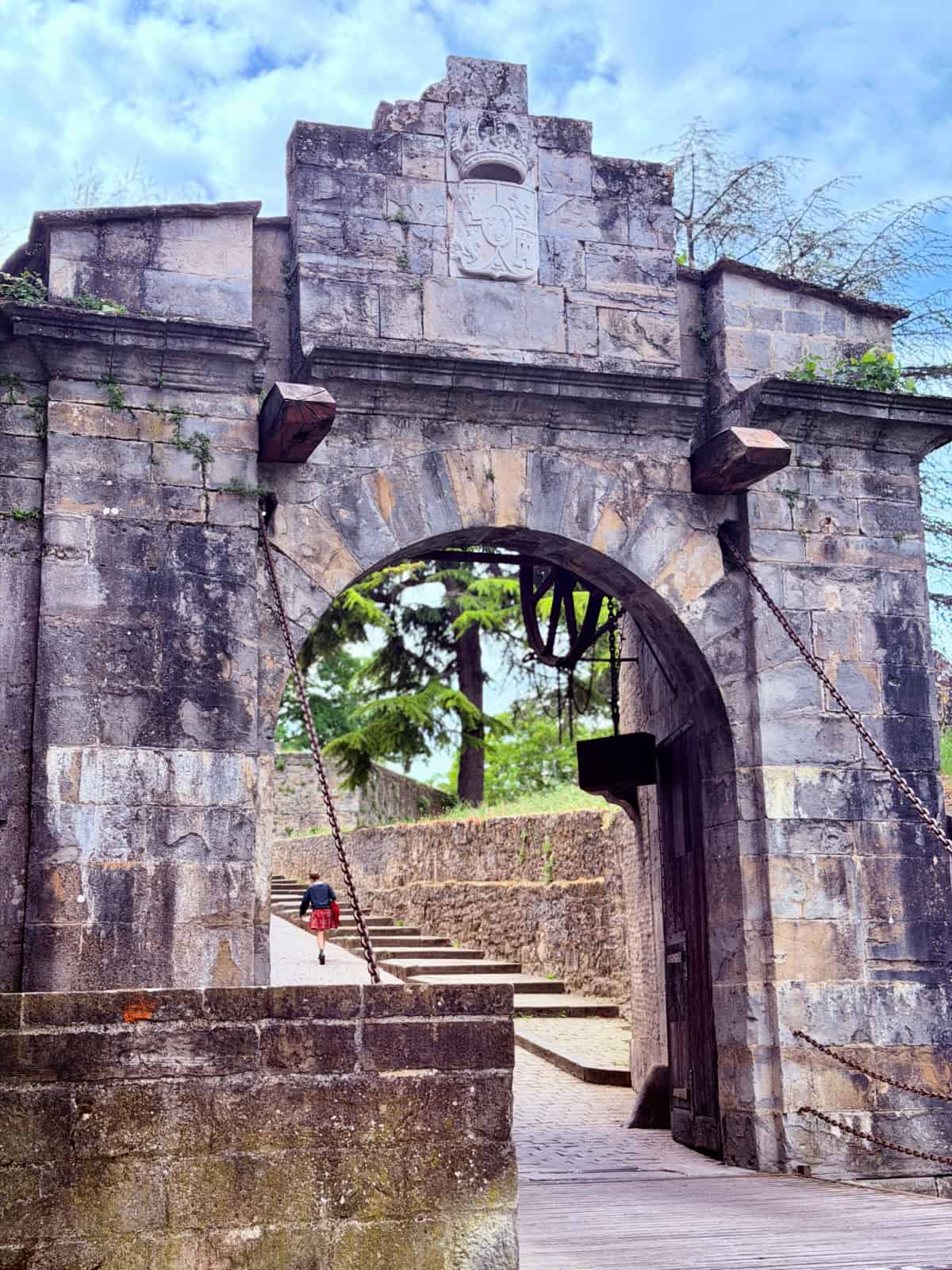 Stone drawbridge entrance to a fortified town on the Camino de Santiago, featuring heavy chains, an arched gateway, and a pilgrim walking uphill beyond the gate.