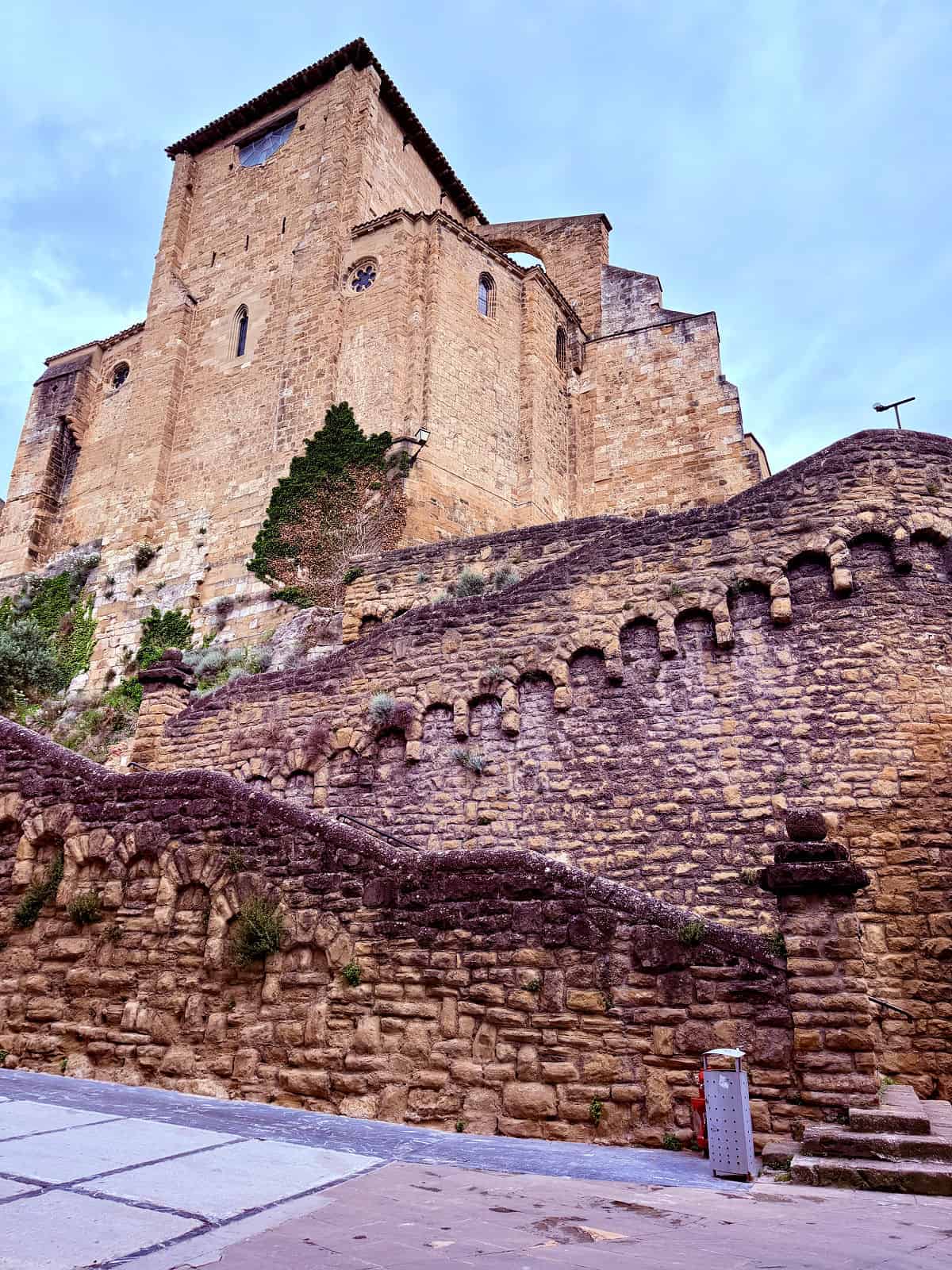 Massive stone church and fortress walls rising above a historic town along the Camino de Santiago under a cloudy sky.