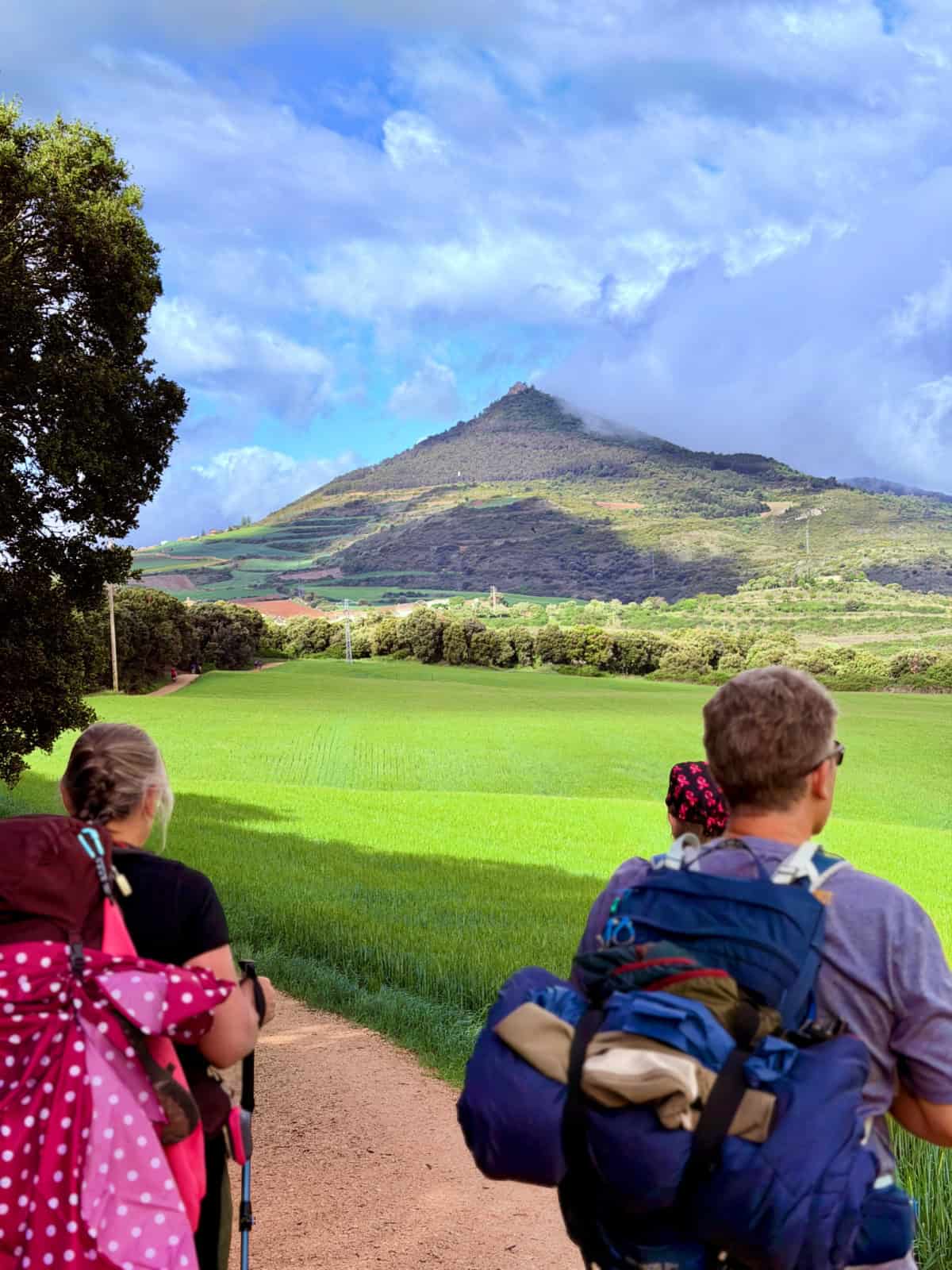 Pilgrims resting along the Camino while looking toward a green mountain peak beneath dramatic clouds and patches of sunlight.