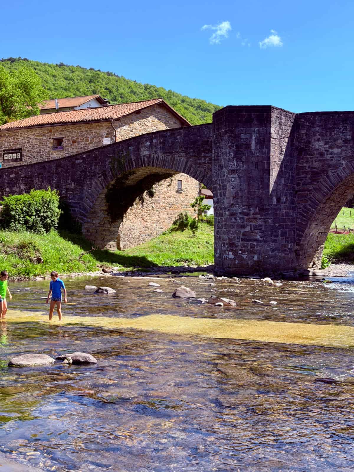 Historic stone bridge spanning a shallow river, with children playing in the water beneath the arches on a sunny day.