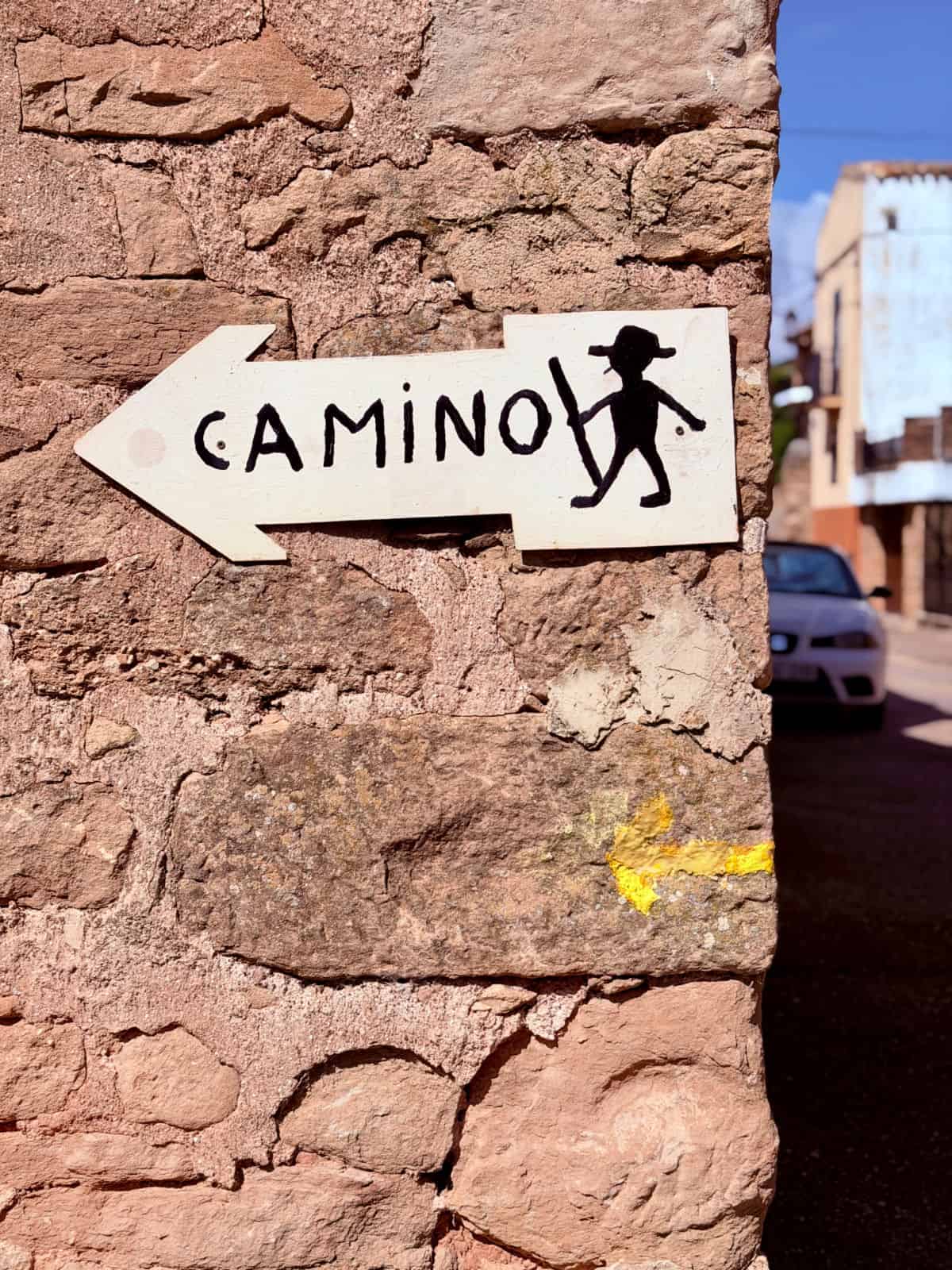 Hand-painted Camino arrow sign mounted on a stone wall, directing pilgrims along the route with a yellow waymark below.