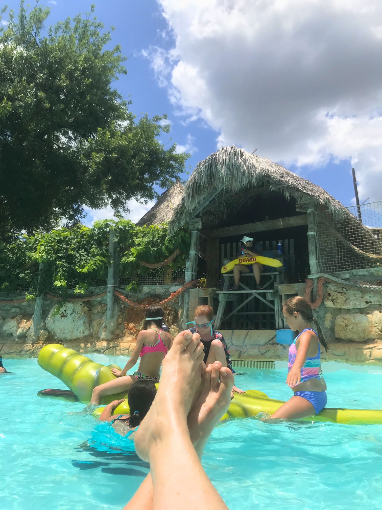 Relaxing view from a pool at Schlitterbahn New Braunfels with children playing on a floating alligator toy, a lifeguard station, and tropical-style thatched-roof structures in the background.