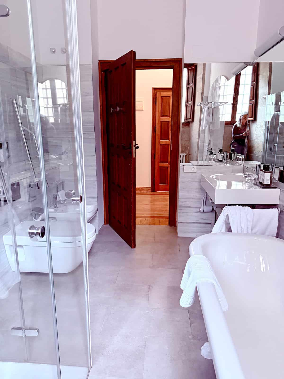 Spacious marble bathroom at the Parador de Santiago de Compostela featuring a glass walk-in shower, freestanding soaking tub, and historic wood accents.