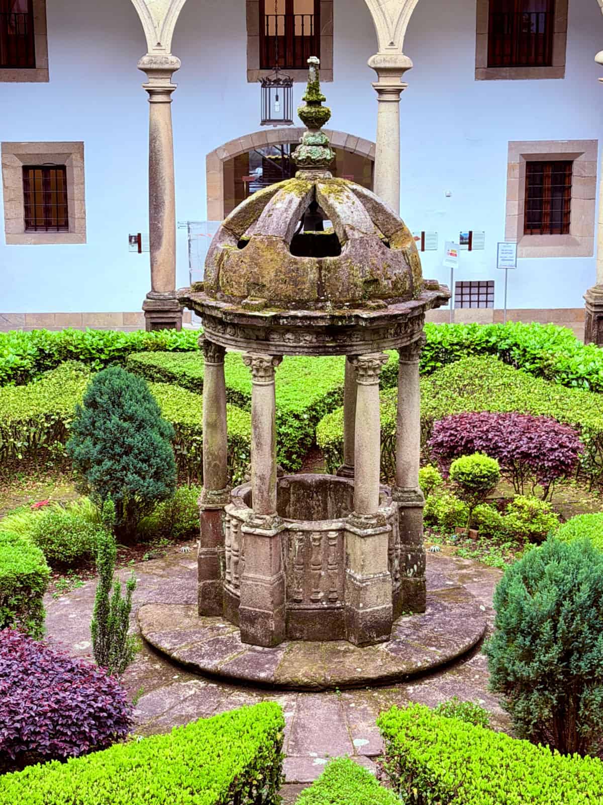 Historic stone fountain beneath a domed canopy in the manicured cloister garden courtyard at the Parador de Santiago.