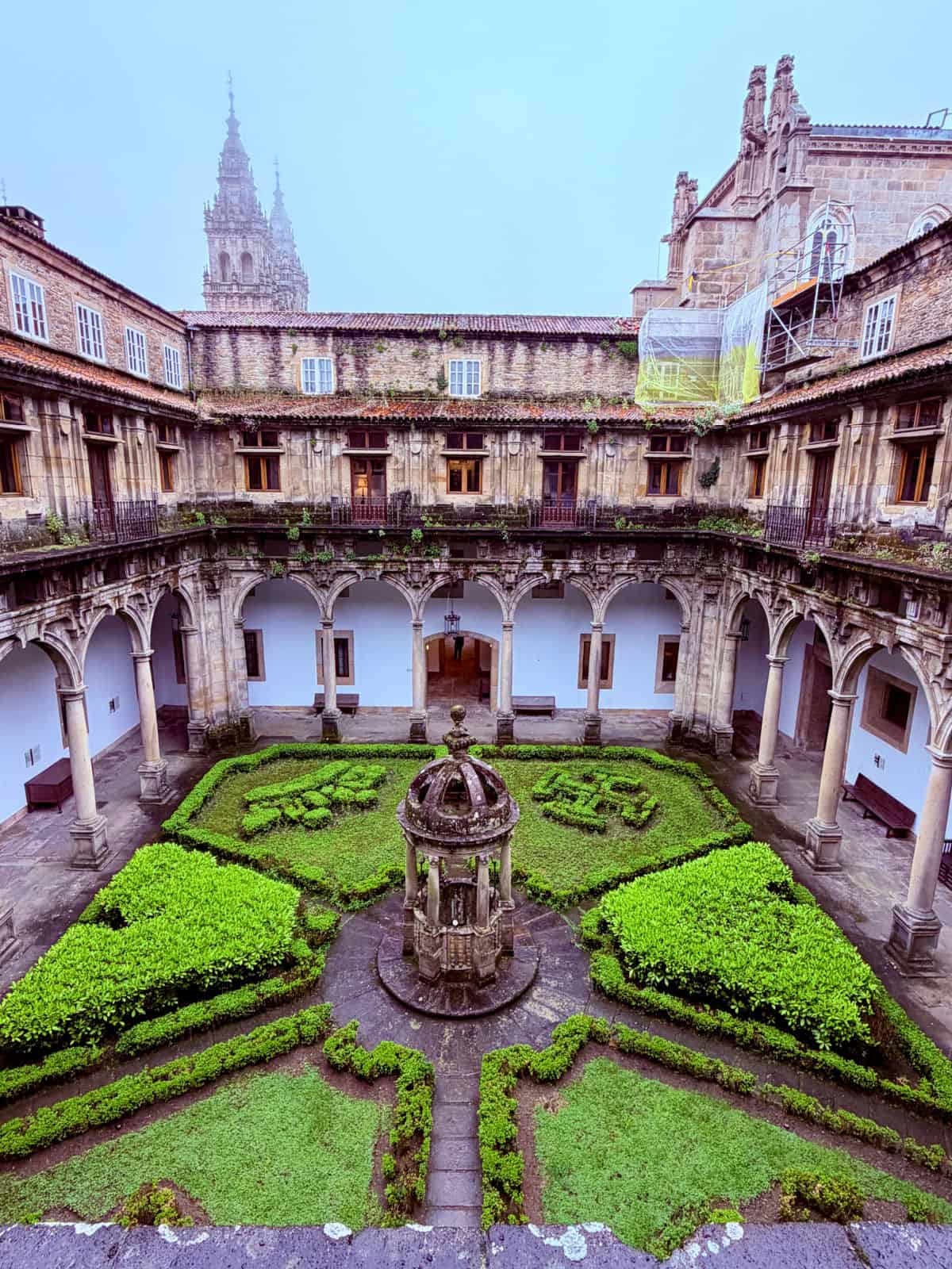Elevated view of the Parador de Santiago’s Renaissance cloister courtyard with trimmed hedges, stone arches, and cathedral towers in the background.