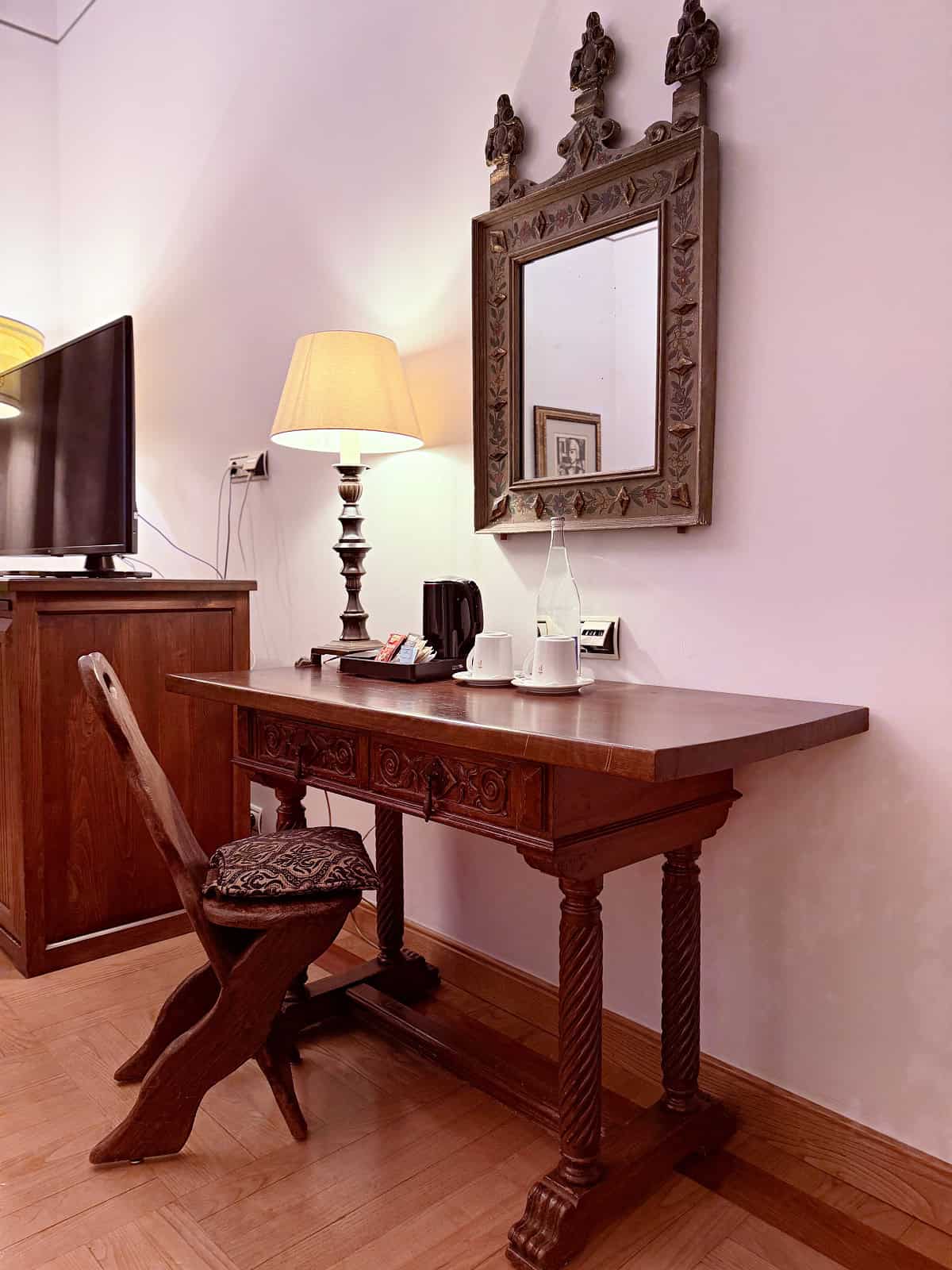 Antique wooden writing desk with carved chair, decorative mirror, and coffee station inside a guest room at the Parador de Santiago.