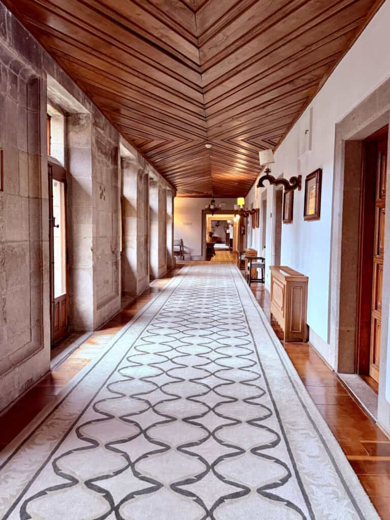 Long historic corridor inside the Parador de Santiago with stone walls, wood-beamed ceiling, and patterned runner carpet.
