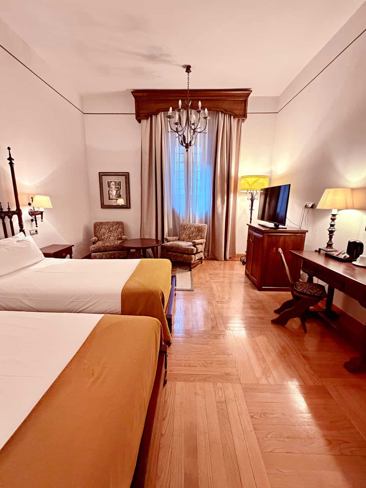 Traditional guest room at the Parador de Santiago featuring twin beds, hardwood floors, antique furnishings, and tall windows.