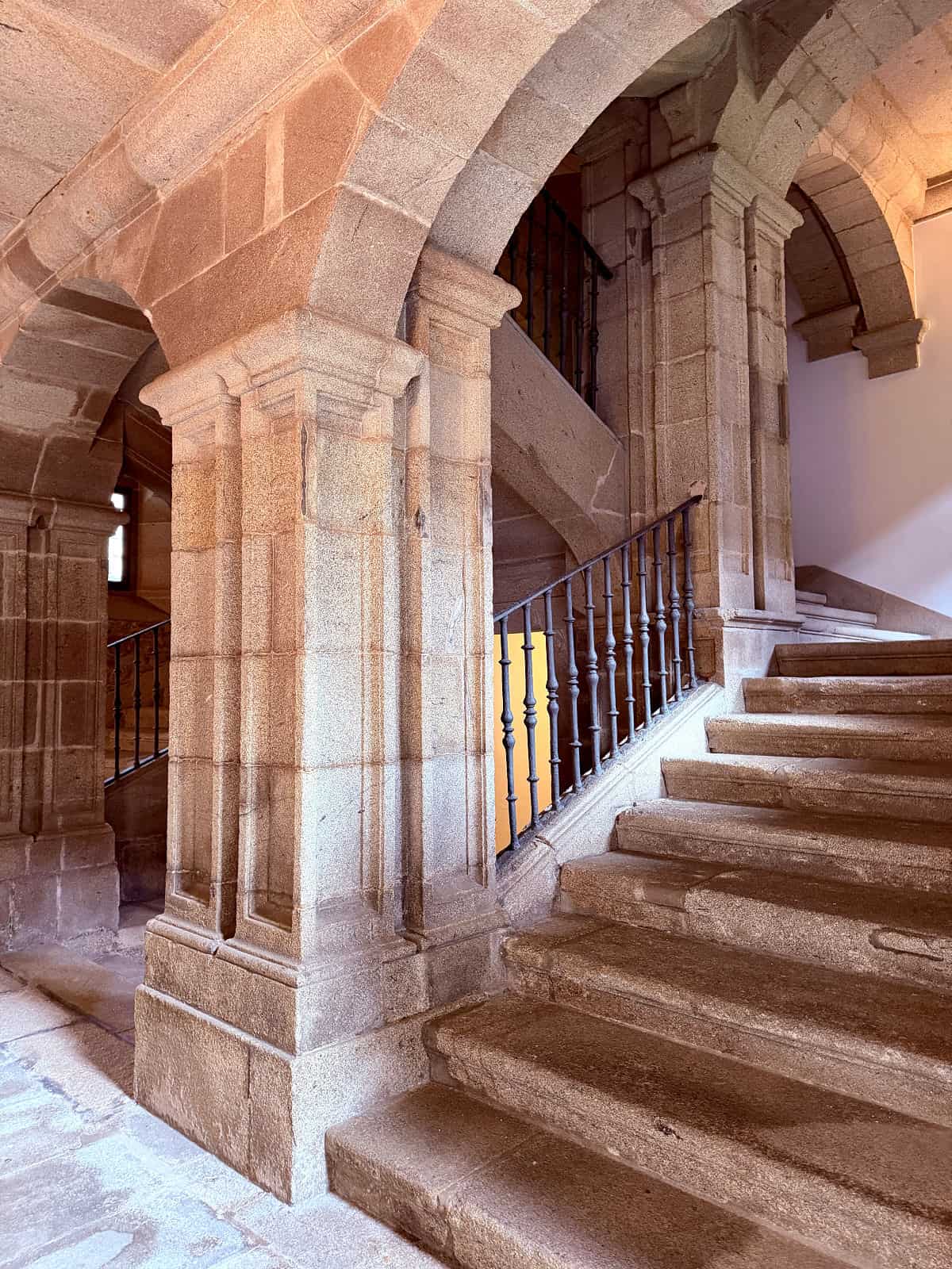 Grand stone staircase with arches and wrought-iron railings inside the historic Parador de Santiago hotel.