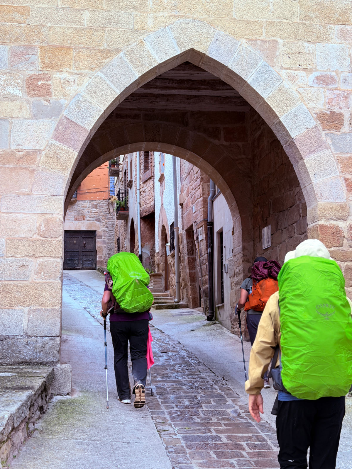 Pilgrims walking through a pointed stone archway onto a narrow medieval street.