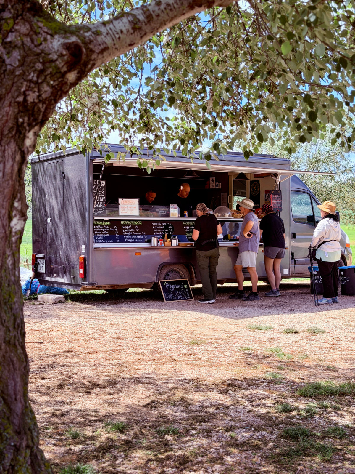 Pilgrims ordering refreshments from a food truck beneath a large shade tree along the Camino.
