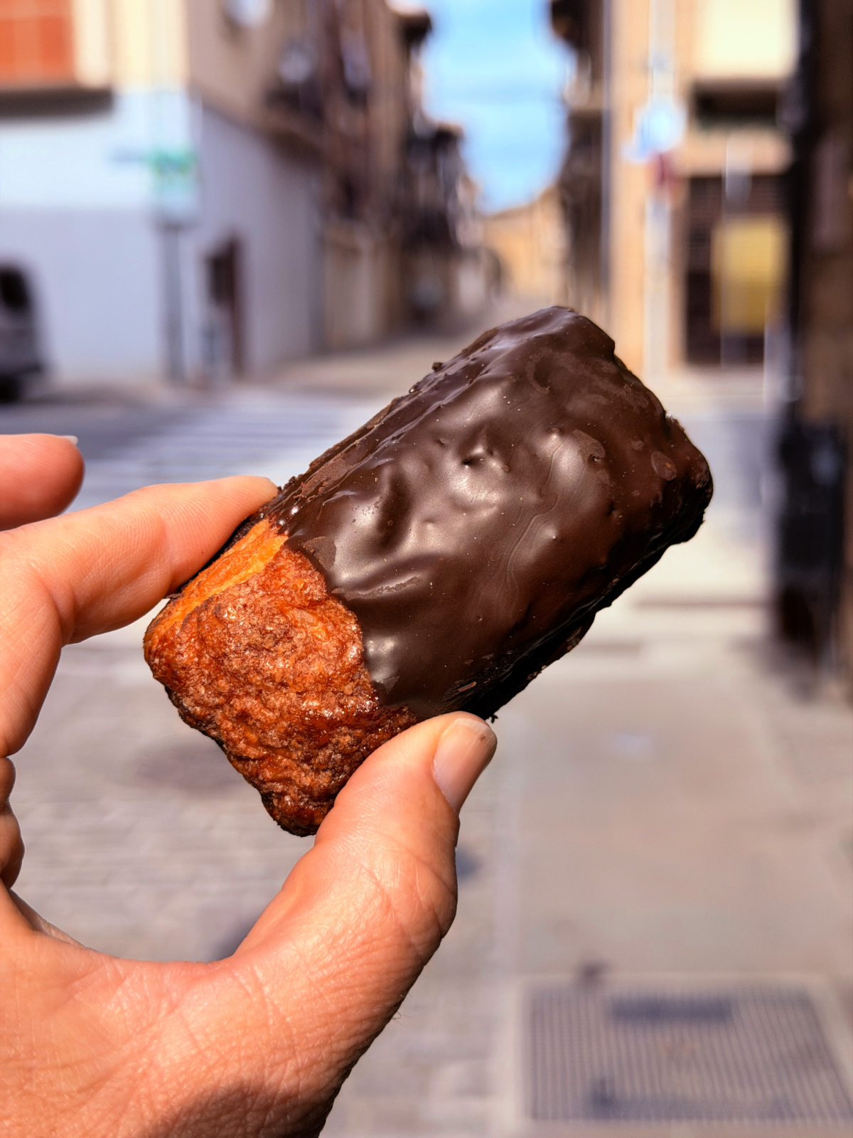Hand holding a small chocolate-covered pastry on a street in Spain.