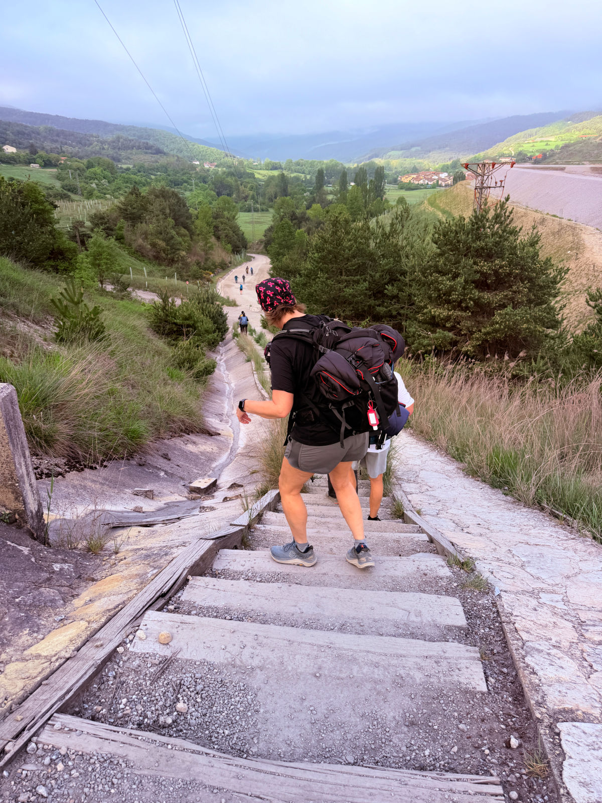 Camino pilgrims carefully descend a steep set of steps overlooking a green valley in Navarra, Spain.