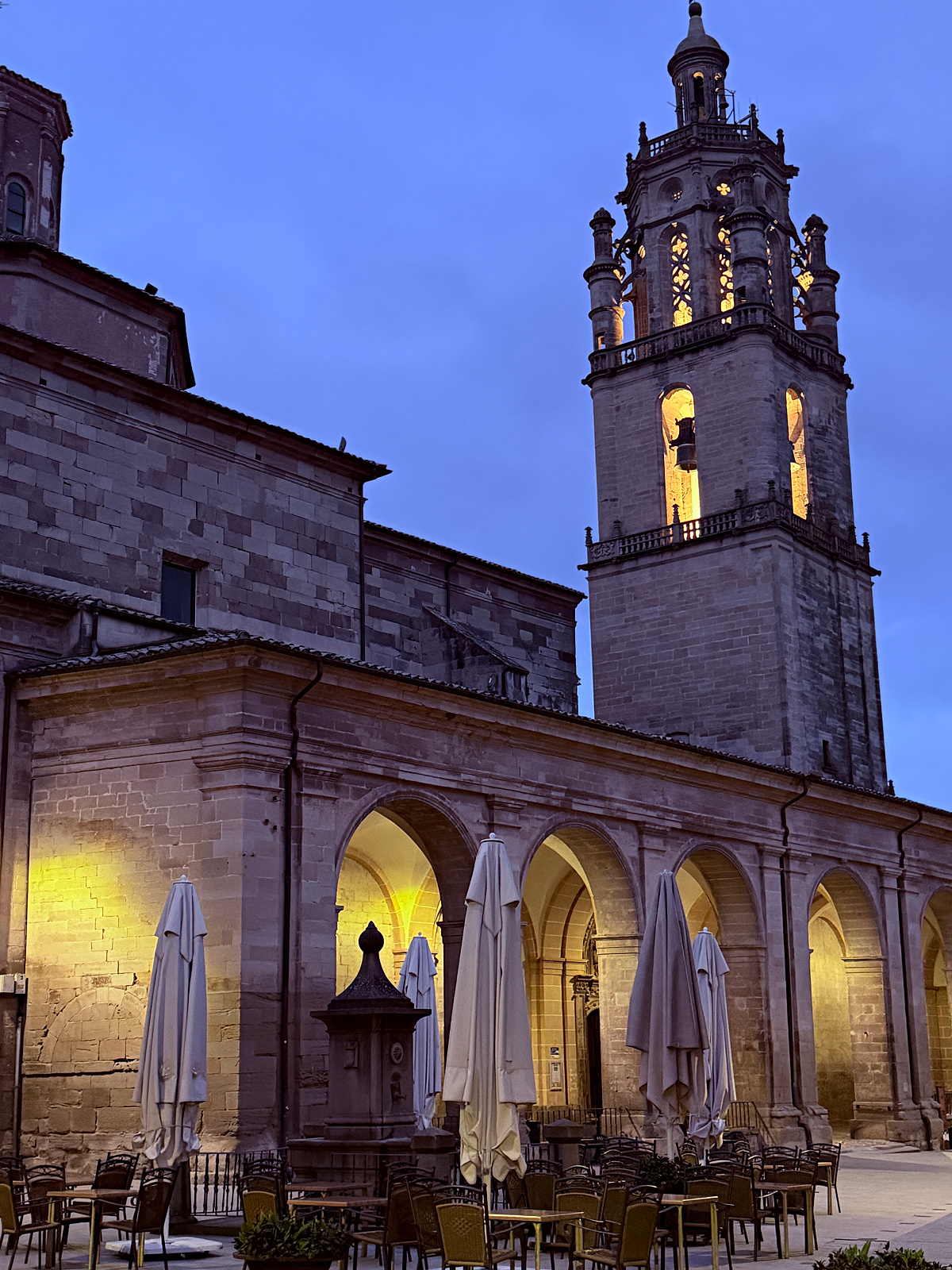 Illuminated church bell tower and empty café tables in Los Arcos at dusk.