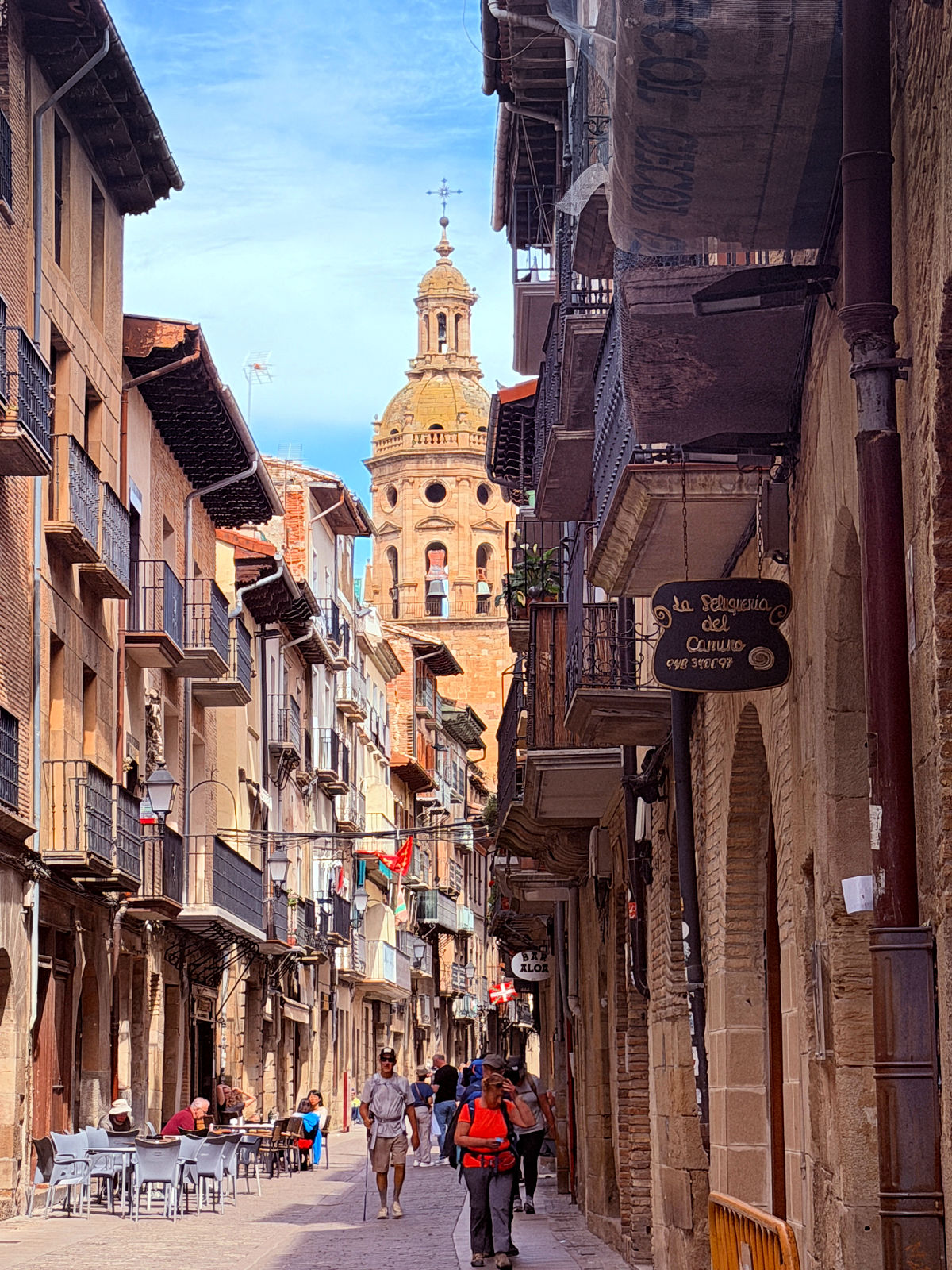 Pilgrims walking along a narrow street toward a church tower in Puente la Reina.