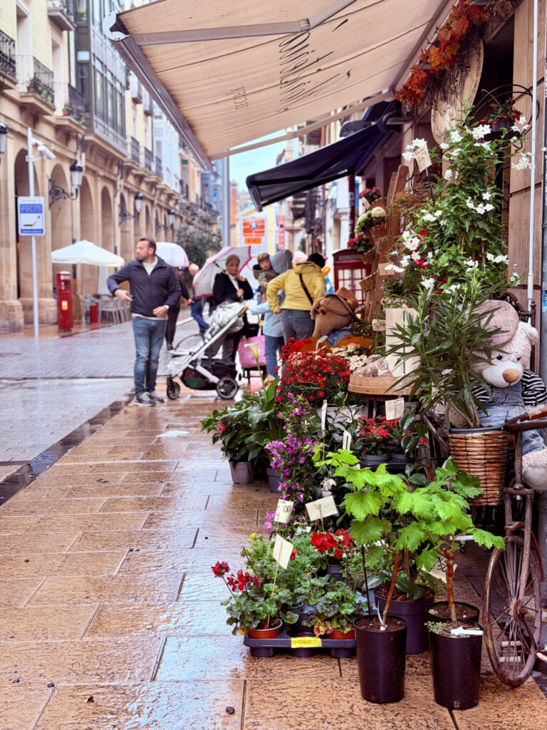 Rain-slicked pedestrian street lined with shops and displays of colorful flowers.