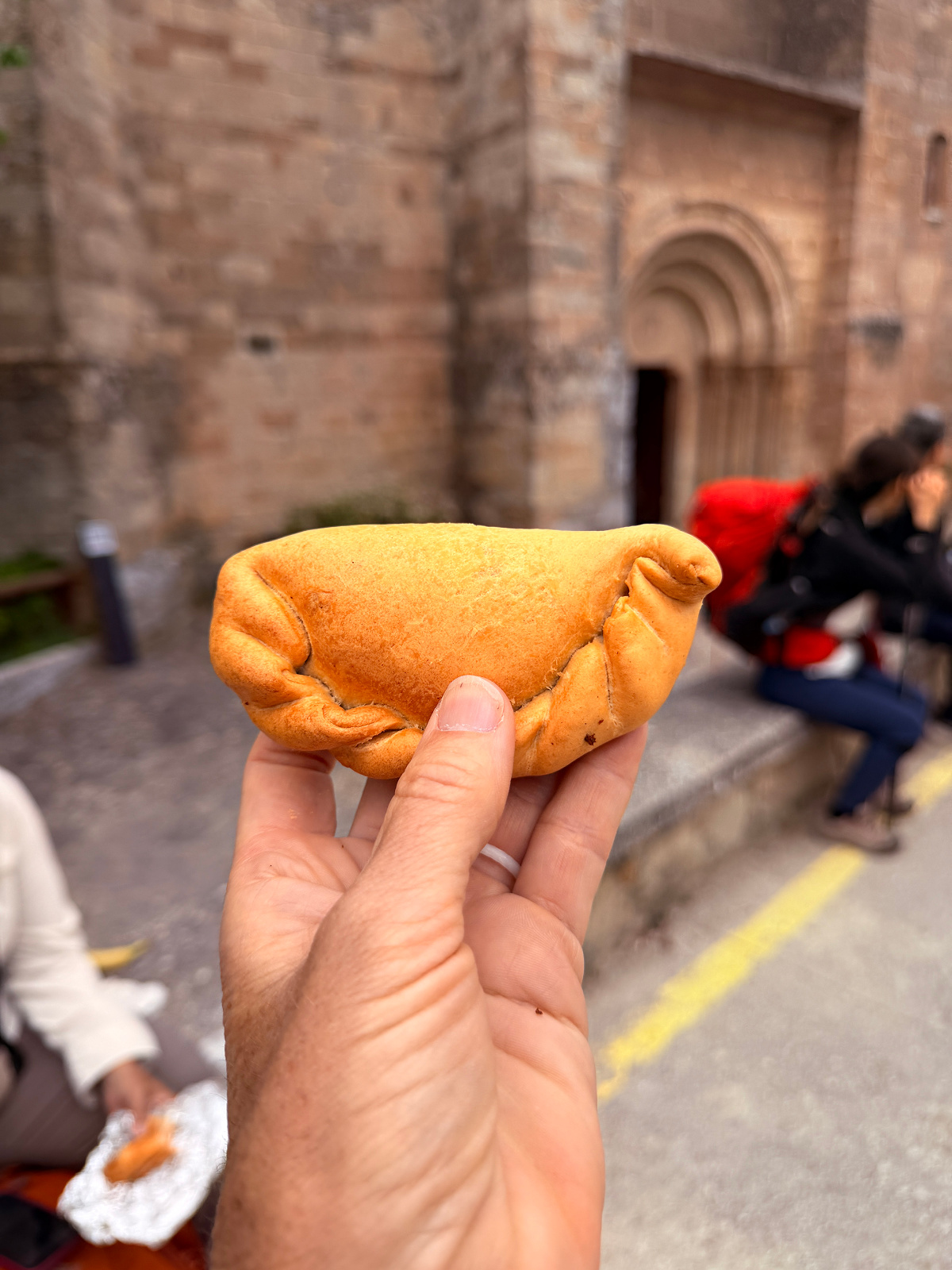 Hand holding a baked Spanish empanada outside a stone church along the Camino de Santiago.