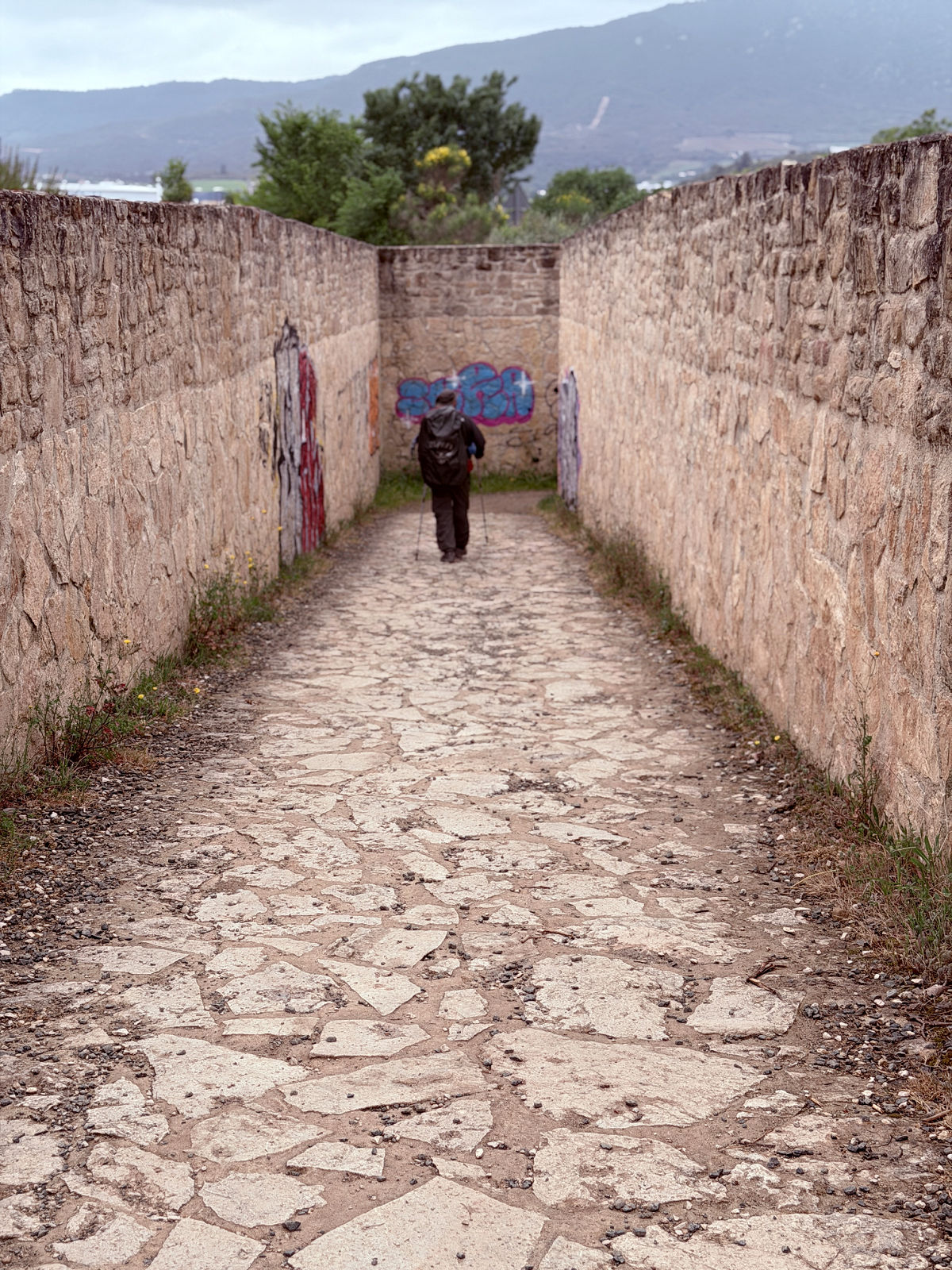 Pilgrim walking along a narrow stone path between high walls.