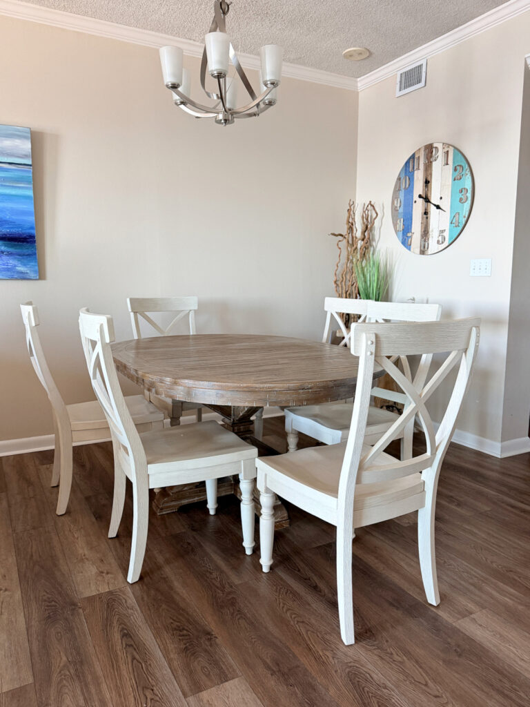Oval wood dining table with six white X-back chairs under a modern chandelier in a beach condo.