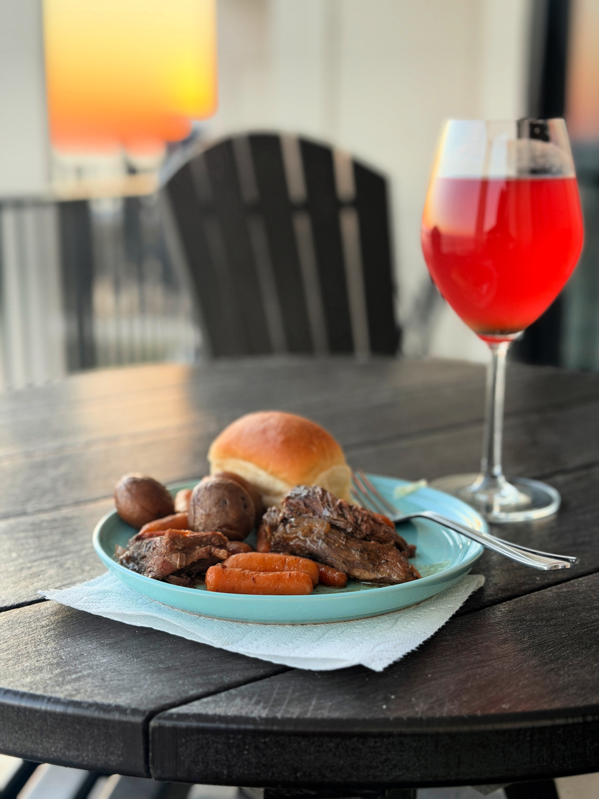 Pot roast with carrots, potatoes, and a dinner roll on a blue plate beside a glass of red drink on a balcony at sunset.