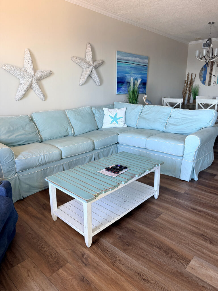 Light blue L-shaped sectional sofa with starfish wall decor and a beachy coffee table in a Phoenix East II condo.