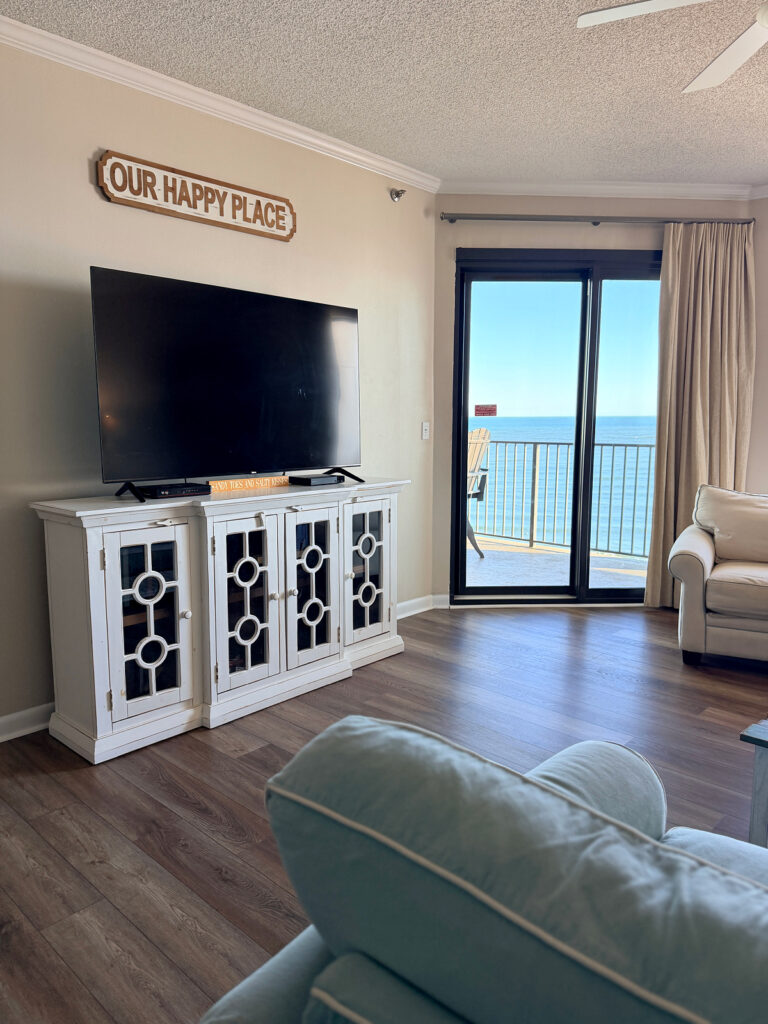 Living room with a large TV on a white console under an "Our Happy Place" sign, with a sliding glass door to a Gulf-view balcony.